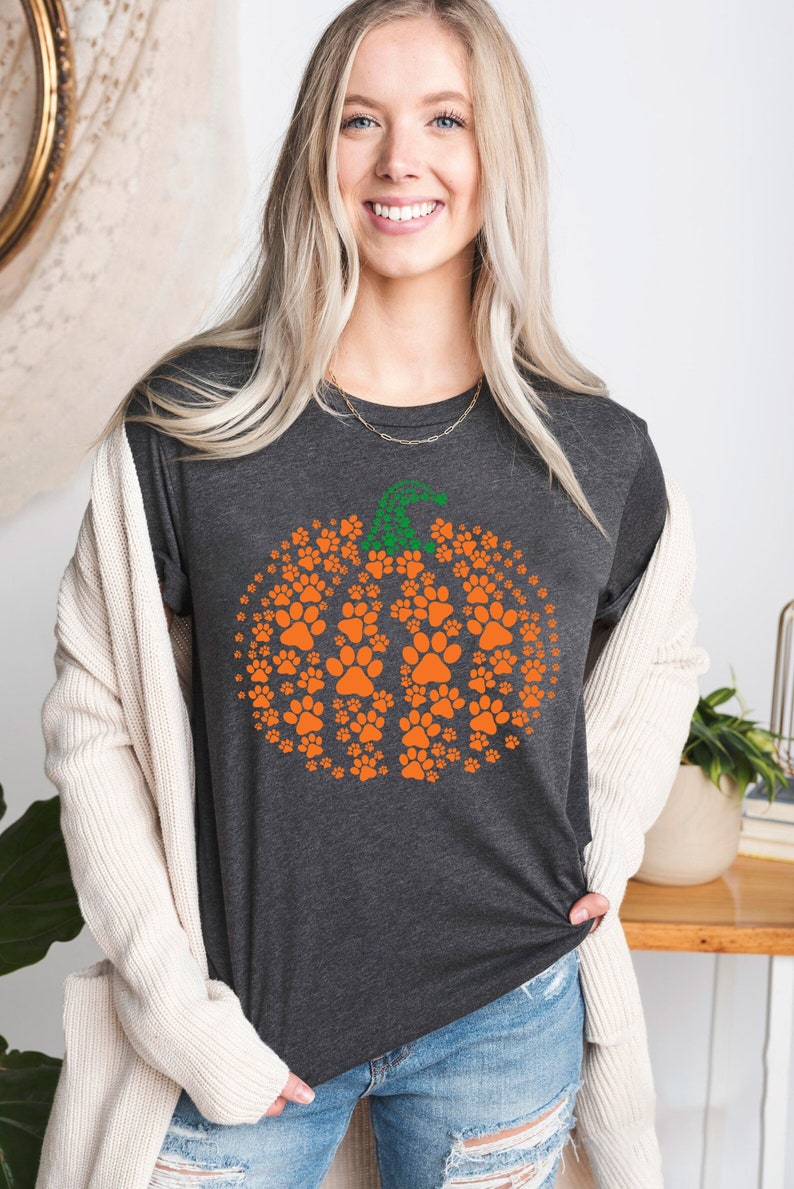 Peut inclure: T-shirt gris fonc&eacute; avec un motif de citrouille en empreintes de pattes orange et une tige verte. La citrouille est compos&eacute;e de nombreuses petites empreintes de pattes. La femme porte le t-shirt et un cardigan cr&egrave;me.