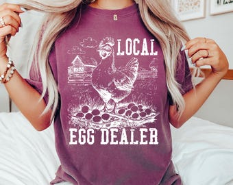 May include: A maroon t-shirt with a white graphic of a hen, eggs, and a barn. The text reads "LOCAL EGG DEALER". The shirt is a comfortable, casual style.