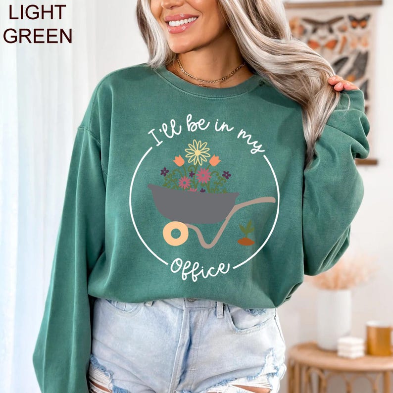 Op de afbeelding: Een lichtgroen sweatshirt met een witte afbeelding van een kruiwagen vol bloemen en de tekst "I'll be in my office".