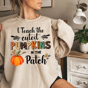 May include: Beige sweatshirt featuring the text "I Teach the cutest PUMPKINS in the Patch." The design incorporates a pumpkin graphic and autumnal leaves. The sweatshirt is a relaxed, casual style.