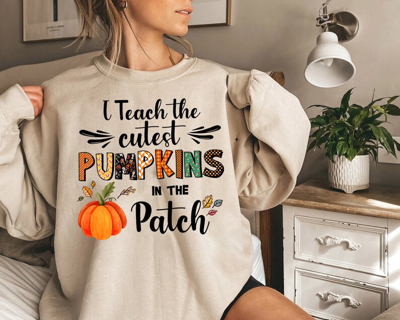 May include: Beige sweatshirt featuring the text "I Teach the cutest PUMPKINS in the Patch." The design incorporates a pumpkin graphic and autumnal leaves. The sweatshirt is a relaxed, casual style.