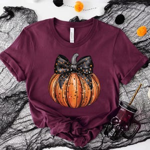 May include: A maroon t-shirt featuring a black and orange pumpkin with a black bow and white stars. The pumpkin is centrally positioned on the shirt.