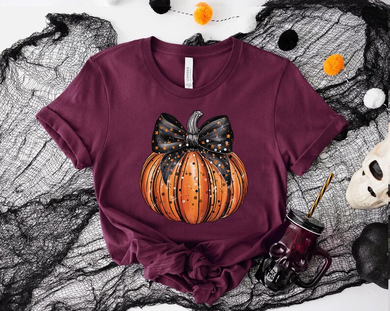 May include: A maroon t-shirt featuring a black and orange pumpkin with a black bow and white stars. The pumpkin is centrally positioned on the shirt.