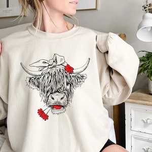 May include: A cream-colored sweatshirt with a black and white illustration of a Highland cow wearing a red flower headband and holding a red flower in its mouth.