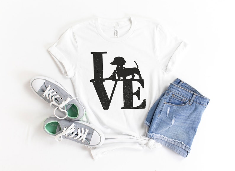 以下が含まれることがあります： 白いTシャツに、黒いシルエットのダックスフントが「LOVE」の文字の中に描かれています。Tシャツは白い表面に置かれ、灰色のスニーカーと青いデニムのショートパンツが置かれています。