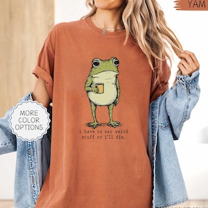 May include: A burnt orange t-shirt featuring a cartoon frog holding a mug. The frog is green with black eyes and a white belly. The text "i have to say weird stuff or i'll die." is below the frog. The shirt has rolled-up sleeves and a relaxed fit.