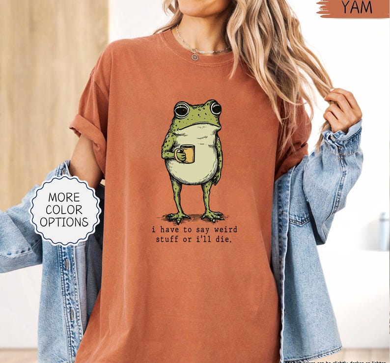 May include: A burnt orange t-shirt featuring a cartoon frog holding a mug. The frog is green with black eyes and a white belly. The text "i have to say weird stuff or i'll die." is below the frog. The shirt has rolled-up sleeves and a relaxed fit.