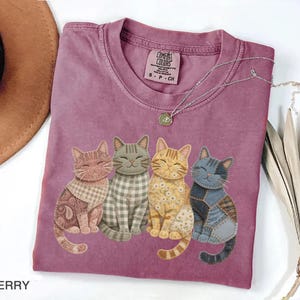 May include: Berry-colored t-shirt featuring four whimsical cat illustrations with patchwork designs. The cats have different patterns and colors, including paisley, gingham, and floral. A silver necklace with a pendant is draped on the shirt.