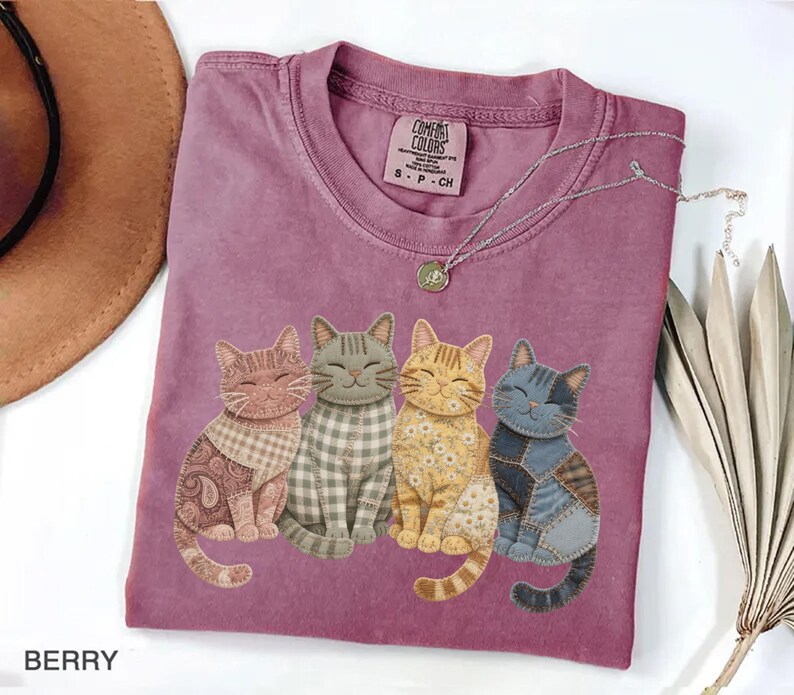 May include: Berry-colored t-shirt featuring four whimsical cat illustrations with patchwork designs. The cats have different patterns and colors, including paisley, gingham, and floral. A silver necklace with a pendant is draped on the shirt.