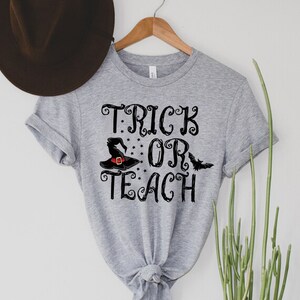 Puede incluir: Camiseta gris claro con las palabras "TRICK OR TEACH" en escritura negra. Un sombrero de bruja negro con una hebilla roja y un gr&aacute;fico de murci&eacute;lago aparecen en el dise&ntilde;o. La camiseta est&aacute; colgada en una percha de madera.