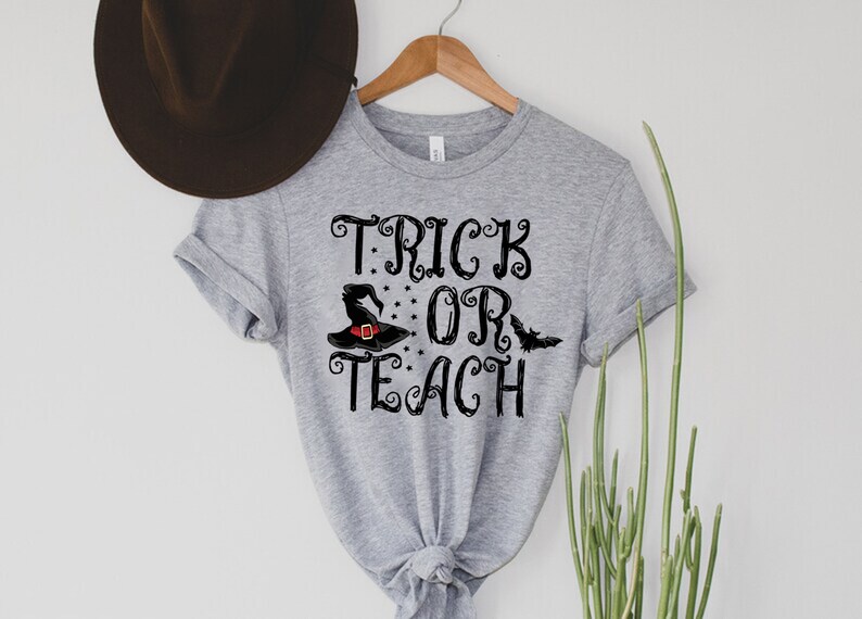 Puede incluir: Camiseta gris claro con las palabras "TRICK OR TEACH" en escritura negra. Un sombrero de bruja negro con una hebilla roja y un gr&aacute;fico de murci&eacute;lago aparecen en el dise&ntilde;o. La camiseta est&aacute; colgada en una percha de madera.