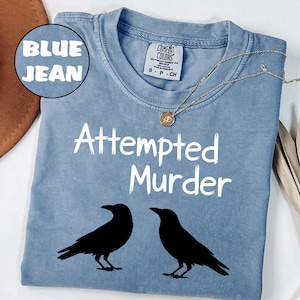 May include: Blue jean colored t-shirt with the words "Attempted Murder" in white, along with two black crow silhouettes. A gold necklace with a pendant is draped across the shirt.