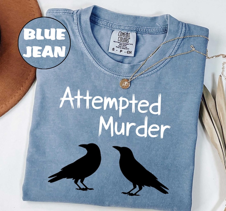 May include: Blue jean colored t-shirt with the words "Attempted Murder" in white, along with two black crow silhouettes. A gold necklace with a pendant is draped across the shirt.