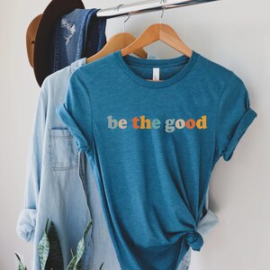 May include: A teal blue t-shirt with the text "be the good" in rainbow colors. The text is in a retro font style.
