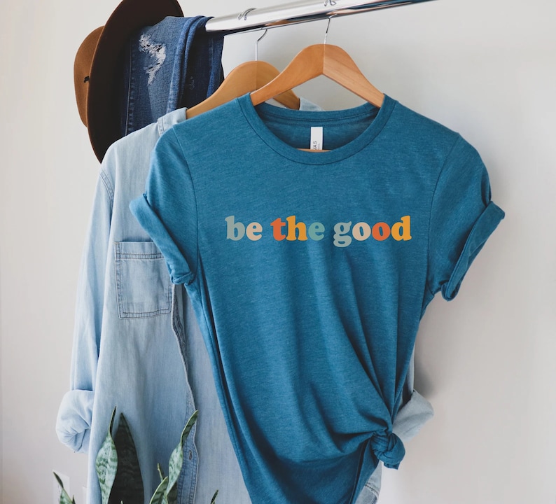 May include: A teal blue t-shirt with the text "be the good" in rainbow colors. The text is in a retro font style.
