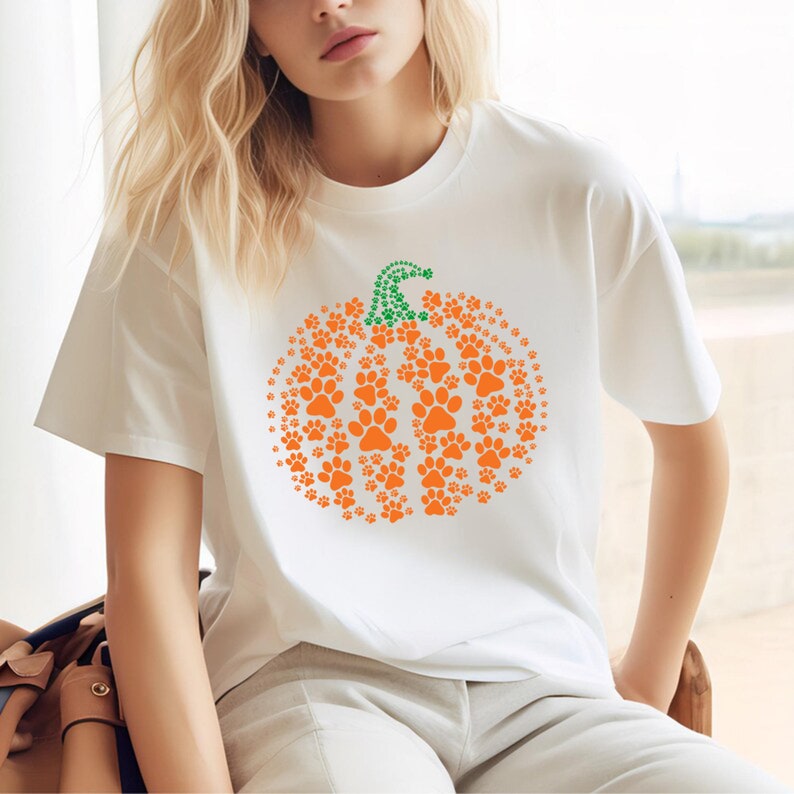 Peut inclure: T-shirt blanc avec un motif de citrouille compos&eacute; d'empreintes de pattes orange et d'une tige verte en pointill&eacute;s. Le t-shirt est de style d&eacute;contract&eacute;, id&eacute;al pour l'automne ou Halloween.