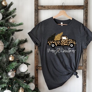 Op de afbeelding: Een donkergrijs T-shirt met een witte afbeelding van een vrachtwagen die een kerstboom vervoert. De vrachtwagen is versierd met een luipaardprint en gouden glitter. De tekst "Merry Christmas" staat onder de vrachtwagen met een hartje.