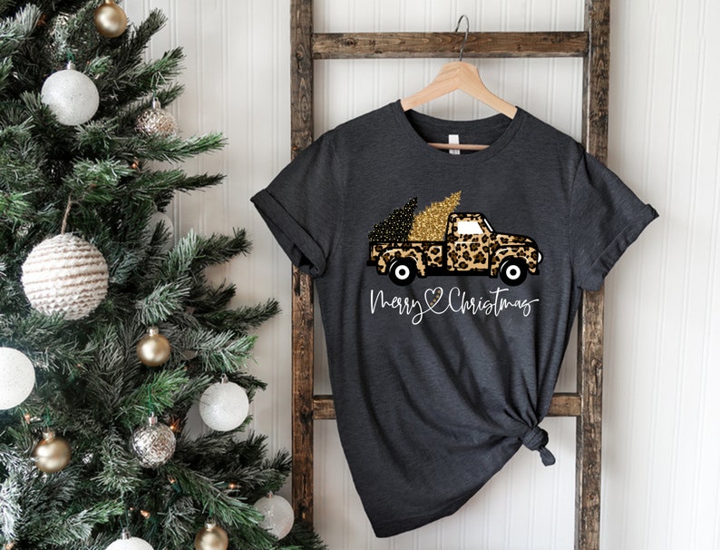 Op de afbeelding: Een donkergrijs T-shirt met een witte afbeelding van een vrachtwagen die een kerstboom vervoert. De vrachtwagen is versierd met een luipaardprint en gouden glitter. De tekst "Merry Christmas" staat onder de vrachtwagen met een hartje.