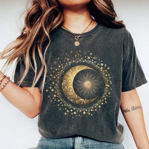 May include: Dark gray t-shirt featuring a gold crescent moon and sun design surrounded by stars. The celestial graphic is centered on the shirt. The shirt has short sleeves and a crew neck.