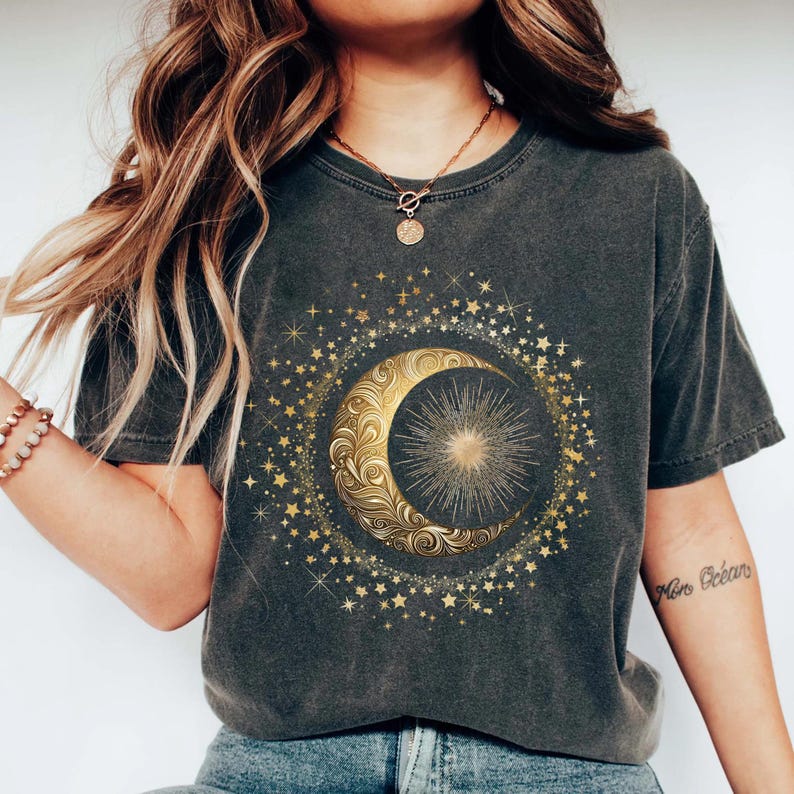 May include: Dark gray t-shirt featuring a gold crescent moon and sun design surrounded by stars. The celestial graphic is centered on the shirt. The shirt has short sleeves and a crew neck.