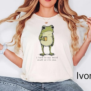 May include: Ivory-colored t-shirt featuring a green frog holding a mug. The frog illustration has a vintage style. The text below the frog reads, "i have to say weird stuff or i'll die."