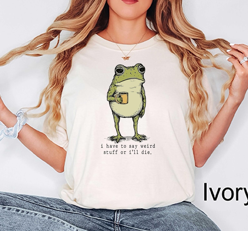 May include: Ivory-colored t-shirt featuring a green frog holding a mug. The frog illustration has a vintage style. The text below the frog reads, "i have to say weird stuff or i'll die."
