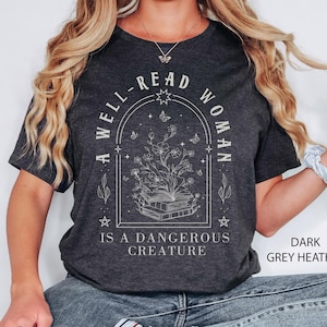 May include: A dark grey heather t-shirt with a white graphic design. The graphic features a book with flowers growing out of it, surrounded by stars and butterflies. The text reads "A well-read woman is a dangerous creature."