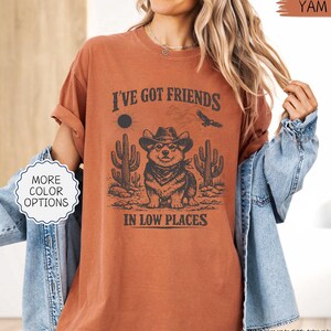 May include: A burnt orange t-shirt with a graphic featuring a cartoon corgi wearing a cowboy hat and bandana, with cacti and the text "I've Got Friends In Low Places". The shirt also has a "More Color Options" button.