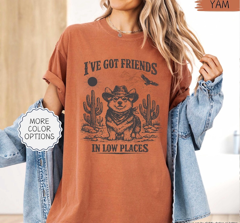 May include: A burnt orange t-shirt with a graphic featuring a cartoon corgi wearing a cowboy hat and bandana, with cacti and the text "I've Got Friends In Low Places". The shirt also has a "More Color Options" button.
