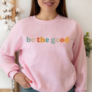 May include: A pink sweatshirt with the text "be the good" in colorful letters.