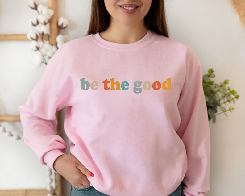 May include: A pink sweatshirt with the text "be the good" in colorful letters.