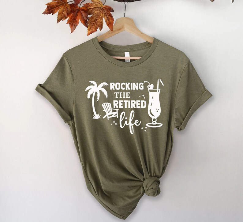 Op de afbeelding: Een groen T-shirt met witte tekst die "Rocking the Retired life" luidt. De tekst wordt vergezeld door een palmboom, een strandstoel en een cocktail.
