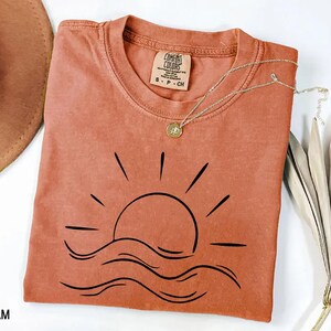 Puede incluir: Una camiseta de color terracota con un gr&aacute;fico negro de un sol poni&eacute;ndose sobre l&iacute;neas onduladas. La camiseta tiene cuello redondo y mangas cortas. Un delicado collar plateado con un colgante descansa sobre la camiseta. La etiqueta dice "Comfort Colors".