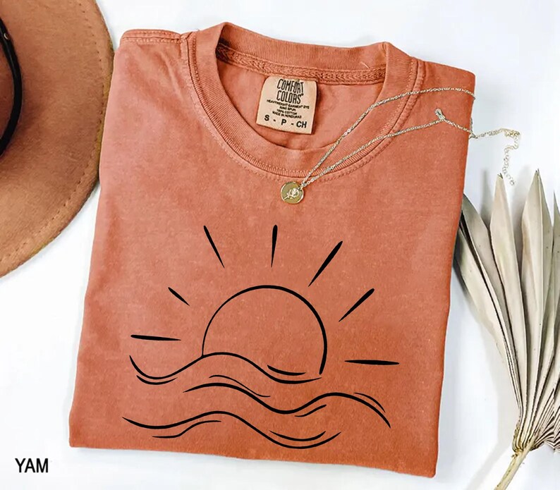 Puede incluir: Una camiseta de color terracota con un gr&aacute;fico negro de un sol poni&eacute;ndose sobre l&iacute;neas onduladas. La camiseta tiene cuello redondo y mangas cortas. Un delicado collar plateado con un colgante descansa sobre la camiseta. La etiqueta dice "Comfort Colors".