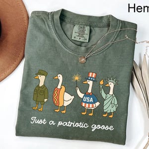 Peut inclure: T-shirt couleur chanvre avec un motif de canard patriotique. Le graphisme présente quatre oies de dessin animé habillées en soldat, en hot-dog, en personnage patriotique avec une étincelle et la Statue de la Liberté. Le texte dit "Juste une oie patriotique."
