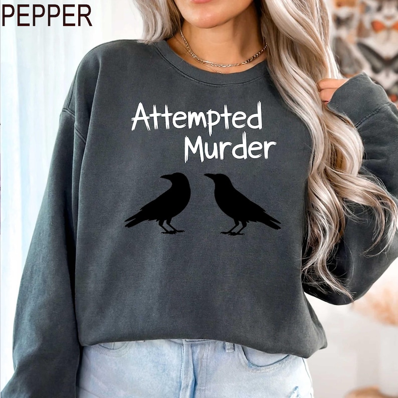 May include: Dark gray sweatshirt with the text "Attempted Murder" in white script above two black crow silhouettes. The sweatshirt has long sleeves and a crew neck.