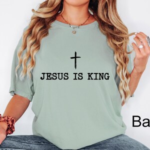 K&ouml;nnte beinhalten: Hellgr&uuml;nes T-Shirt mit dem schwarzen Text &bdquo;JESUS IS KING&ldquo; und einem schwarzen Kreuz dar&uuml;ber. Das Shirt ist einfarbig und hat eine lockere Passform.