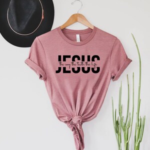 Pu&ograve; includere: Una maglietta color rosa polvere con la parola "JESUS" in lettere maiuscole nere e la frase "the way the truth the life" in scrittura corsiva nera.