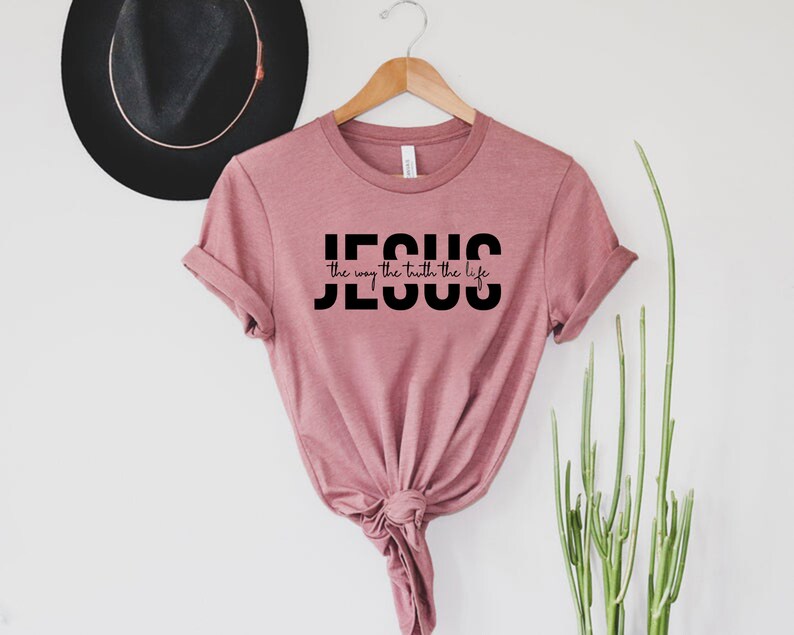 Pu&ograve; includere: Una maglietta color rosa polvere con la parola "JESUS" in lettere maiuscole nere e la frase "the way the truth the life" in scrittura corsiva nera.