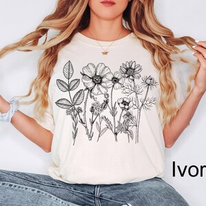 May include: Ivory-colored t-shirt featuring a detailed black and white floral illustration. The design includes various botanical elements, such as flowers and leaves. The shirt is paired with blue jeans.