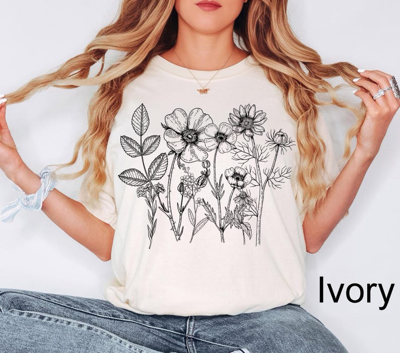May include: Ivory-colored t-shirt featuring a detailed black and white floral illustration. The design includes various botanical elements, such as flowers and leaves. The shirt is paired with blue jeans.