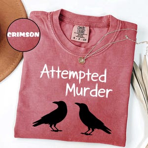 May include: Crimson-colored t-shirt with the text "Attempted Murder" and two black raven silhouettes. The shirt has a crew neck and a small tag at the neckline. A silver necklace with a pendant is draped across the shirt.