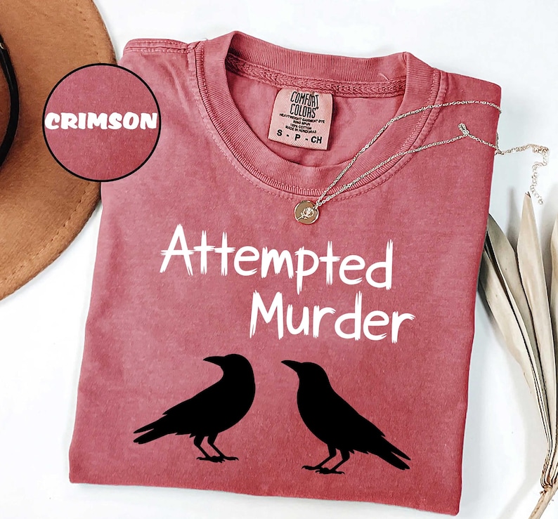 May include: Crimson-colored t-shirt with the text "Attempted Murder" and two black raven silhouettes. The shirt has a crew neck and a small tag at the neckline. A silver necklace with a pendant is draped across the shirt.