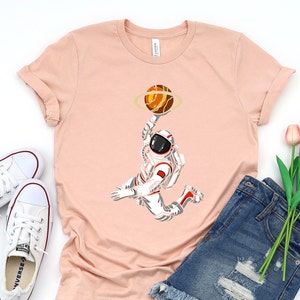 Può includere: Una maglietta rosa con una grafica di un astronauta che schiaccia una palla da basket, che è un pianeta con anelli. L'astronauta indossa una tuta spaziale bianca con una striscia rossa e un casco blu.