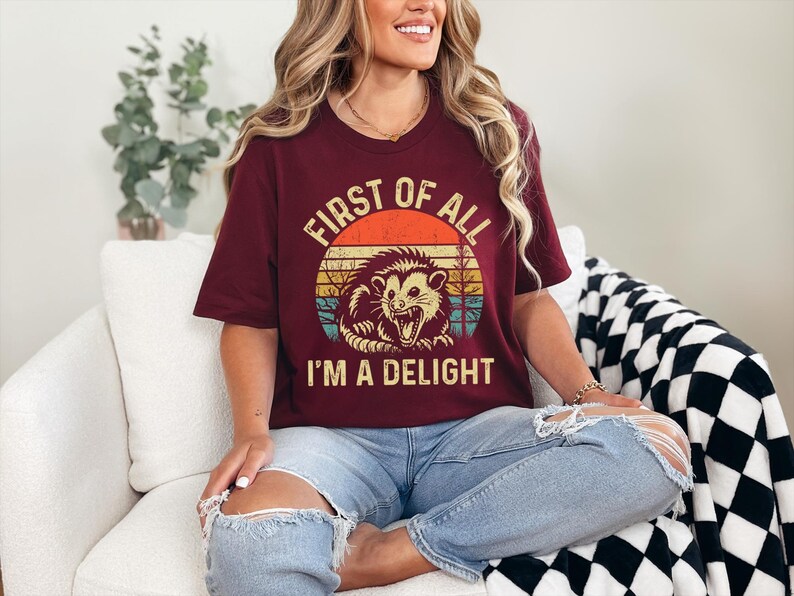 May include: A maroon t-shirt with a vintage-style graphic of an opossum with the text "First of all I'm a delight".