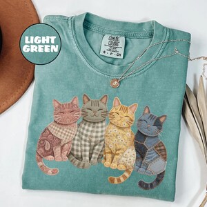 May include: Light green Comfort Colors t-shirt featuring four smiling cat illustrations with patchwork patterns. The cats are in shades of brown, gray, and yellow. A silver necklace with a small pendant is also visible.