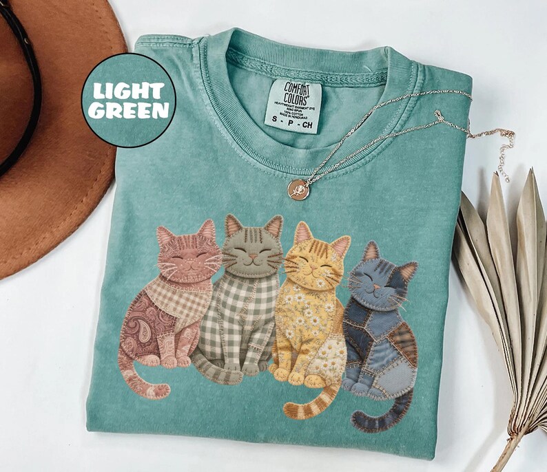 May include: Light green Comfort Colors t-shirt featuring four smiling cat illustrations with patchwork patterns. The cats are in shades of brown, gray, and yellow. A silver necklace with a small pendant is also visible.