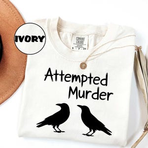 May include: Ivory-colored t-shirt with the text "Attempted Murder" and two black crow silhouettes. The shirt has a Comfort Colors label and a gold necklace. The word "IVORY" is in a circle.