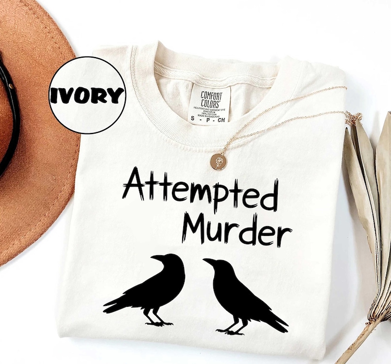 May include: Ivory-colored t-shirt with the text "Attempted Murder" and two black crow silhouettes. The shirt has a Comfort Colors label and a gold necklace. The word "IVORY" is in a circle.