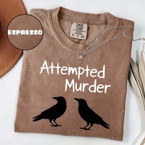 May include: Espresso-colored t-shirt with the words "Attempted Murder" in white, along with two black crow silhouettes. The shirt has a crew neck and short sleeves. A silver necklace with a pendant is draped across the shirt.